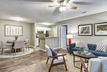 Staged living room with blue couch, two gray accent chairs, ceiling fan, decorative rug, wall art, dining room with table and four chairs, and kitchen in the background.